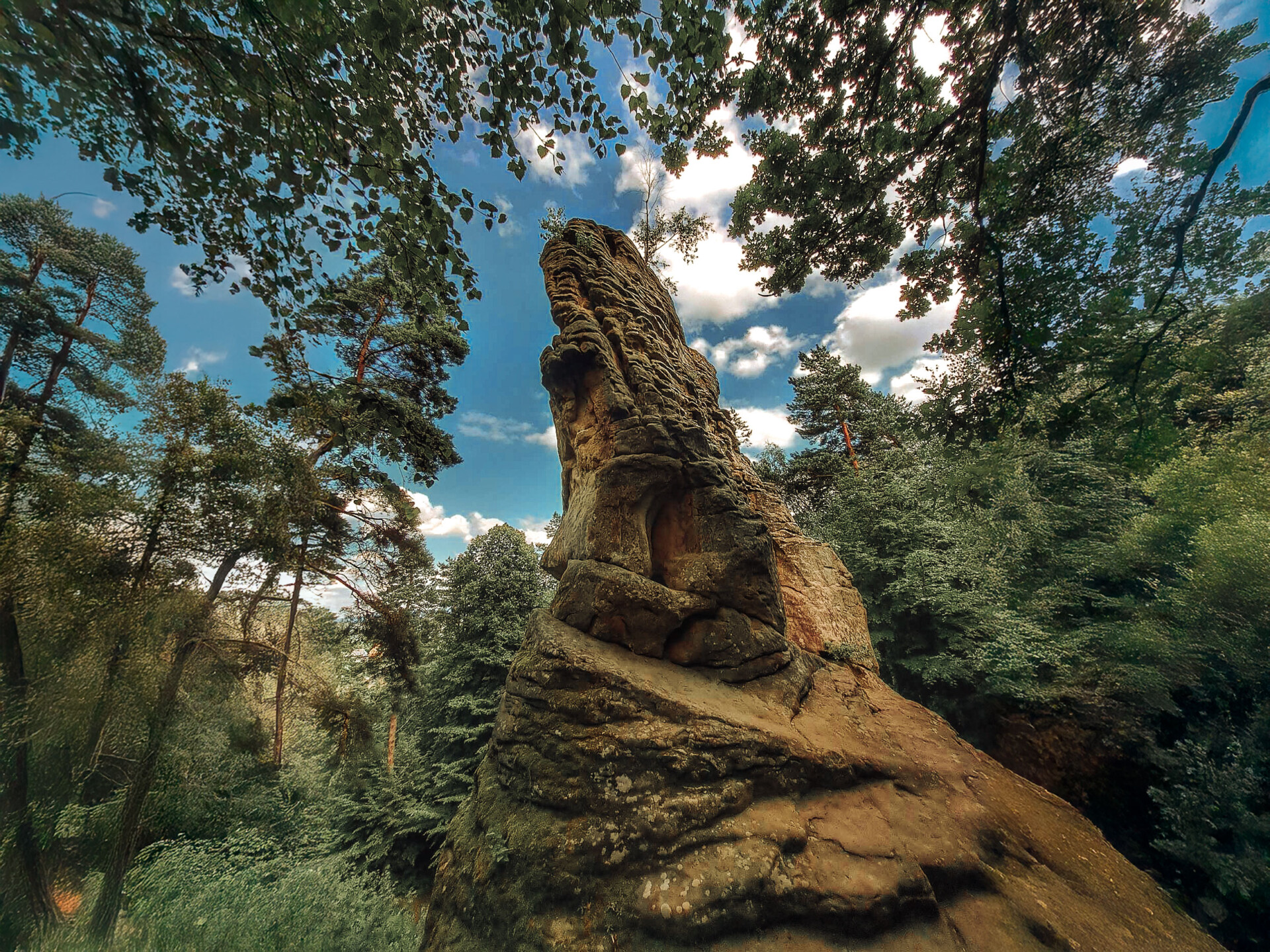 Potężna formacja skalna Kamień Leski – Bieszczadska Legenda w otoczeniu gęstego, zielonego lasu, widziana z dołu przez liście drzew, na tle niebieskiego nieba z chmurami